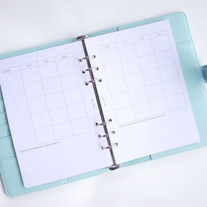 May include: A white weekly planner with a light blue cover. The planner has a section for priorities, a daily schedule, and a section for self-care reminders. The text "This month's goals (personal & professional)" is at the bottom of the page.