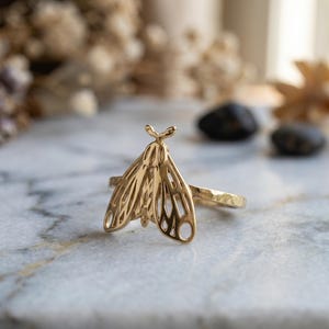 May include: A gold-colored ring featuring a detailed moth design. The ring's band has a textured finish, and the moth's wings are intricately cut out. The ring is set against a blurred background with natural elements.