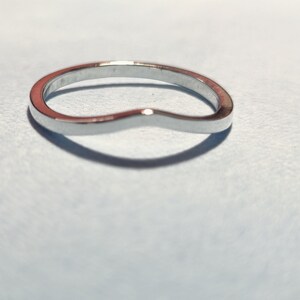 May include: A curved, silver-colored ring with a smooth, polished surface. The ring has a gentle wave design, creating a subtle, elegant look. The ring is set against a light blue background.