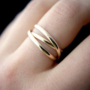 Könnte beinhalten: Nahaufnahme eines goldenen Rings mit drei Bändern, die sich um den Finger legen. Der Ring hat eine polierte, reflektierende Oberfläche, und die Bänder sind glatt und abgerundet. Ein schlichtes, elegantes Schmuckstück.