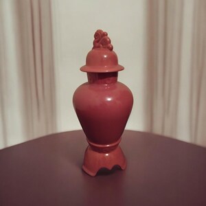 May include: A red ceramic jar with a lid and a decorative top. The jar has a bulbous body and a flared base. The lid is topped with a small, ornate design. The jar is displayed on a dark surface.