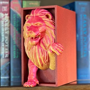 May include: A red and gold lion bookend. The lion's head and paws extend from a red rectangular box. The lion has a detailed mane and facial features. The bookend is designed to hold books on a shelf.