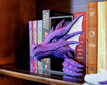 May include: A purple dragon head bookend with blue eyes and white accents. The dragon is positioned between books on a shelf. The books have various colors and titles, including "Rebecca Yarros" and "David Day".
