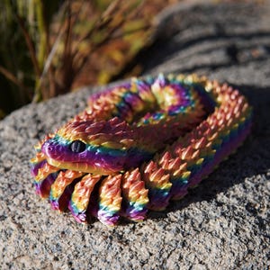 Peut inclure: Une figurine de serpent articulée et enroulée présentant un dégradé arc-en-ciel vibrant de couleurs métalliques, notamment violet, bleu, jaune et orange. Le serpent possède une surface texturée en écailles avec des segments pointus et des yeux sombres.