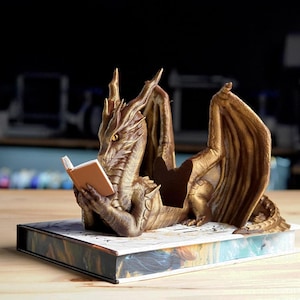 May include: A bronze-colored dragon figurine reading a small book. The dragon is resting on a decorative book with colorful illustrations. The dragon has detailed wings and scales, and the book has a tan cover.