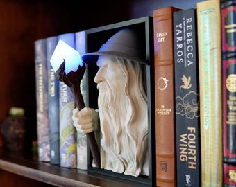 May include: A bookshelf with a decorative sculpture of a wizard holding a staff with a glowing blue crystal. The sculpture is set within a black frame, and the books are in the background. The books have various colors and titles.