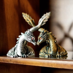 May include: Two dragon figurines with detailed scales and red eyes face each other. One is silver, the other bronze, and they are positioned on a wooden shelf. A decorative element with leaf-like details is above them.
