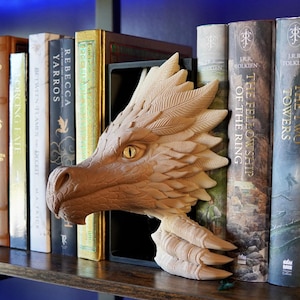 May include: A detailed, light brown dragon head bookend with a yellow eye, placed among a row of books on a wooden shelf. The bookend is made of a carved material and adds a fantasy theme to the bookshelf.