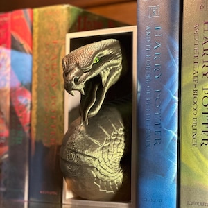 Könnte beinhalten: Eine detaillierte grüne Schlangenskulptur, mit sichtbarem Kopf und offenem Maul, erscheint aus einem weißen Rahmen zwischen zwei "Harry Potter" Büchern. Die Bücher sind in Blau- und Grüntönen gehalten. Die Skulptur hat grüne Augen.