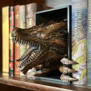 May include: A detailed, brown dragon bookend with sharp teeth and textured scales. The dragon's head emerges from a black rectangular frame, with its claws holding the books. The bookend is placed among a collection of books.