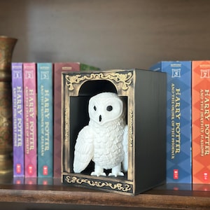 Puede incluir: Una figura de búho blanco dentro de una caja decorativa negra y dorada con detalles ornamentados. La caja está sobre un estante con una fila de libros de Harry Potter en el fondo. Los libros tienen títulos como "Harry Potter y las Reliquias de la Muerte".