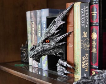 May include: A detailed dragon bookend in silver and black, with red eyes, positioned between books on a shelf. The dragon's head and clawed hand emerge from the bookend, adding a fantasy element to the bookshelf.