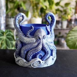 May include: A blue and silver octopus-themed planter. The planter features an octopus design with tentacles forming handles and a detailed octopus face. The base has a wave pattern. The planter is likely made of resin or ceramic.