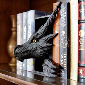 May include: A black dragon bookend with a detailed, textured design. The dragon's head and claw emerge from behind a row of books, with a striking yellow eye. The bookend adds a fantasy theme to a bookshelf.
