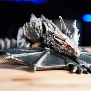 Puede incluir: Figura de dragón plateado con ojos rojos y alas extendidas. El dragón está posado sobre una superficie de madera, con la cola enroscada detrás. La figura presenta detalles intrincados.