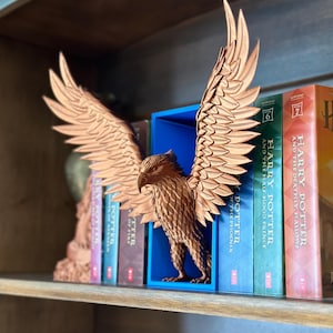 Könnte beinhalten: Ein hölzernes Adler-Buchstütze mit ausgebreiteten Flügeln, in brauner Ausführung, steht zwischen Büchern auf einem Regal. Die Buchstütze befindet sich in einer blauen Box. Die Bücher tragen die Titel "Harry Potter und die Heiligtümer des Todes", "Harry Potter und der Halbblutprinz" und "Harry Potter und der Orden des Phönix".