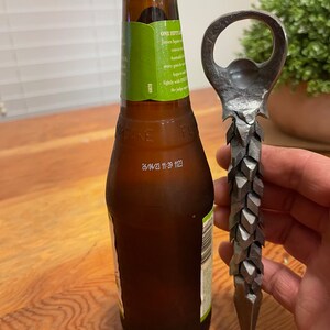 May include: A close-up of a bottle opener with a unique, spiky design. The opener is made of metal and has a sharp point at the bottom. The bottle opener is next to a brown glass bottle of beer.