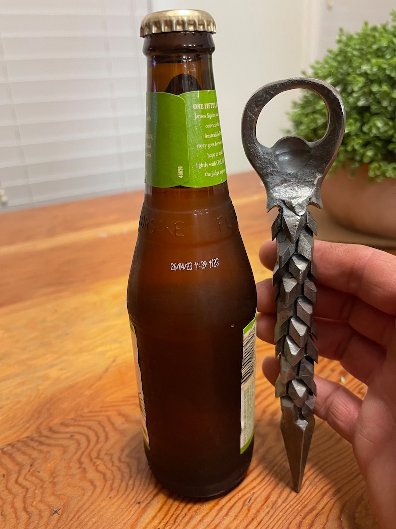 May include: A close-up of a bottle opener with a unique, spiky design. The opener is made of metal and has a sharp point at the bottom. The bottle opener is next to a brown glass bottle of beer.