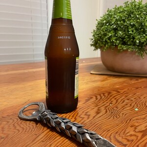 May include: A silver bottle opener with a textured, scaly design. The opener is lying on a wooden surface next to a brown glass bottle.