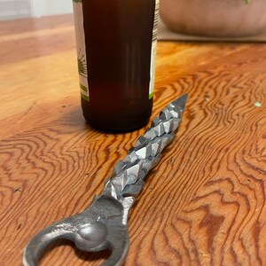 May include: A silver bottle opener with a textured, jagged handle. The opener is lying on a wooden surface.