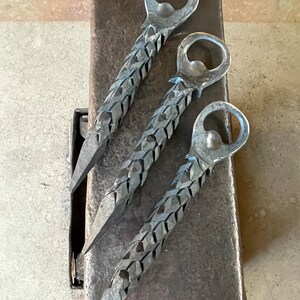 May include: Three silver metal bottle openers with a braided design. The bottle openers are lying on a gray metal surface.