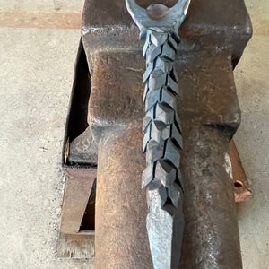 May include: A metal bottle opener with a unique, textured design. The opener is shaped like a spear with a ring at the top for opening bottles.