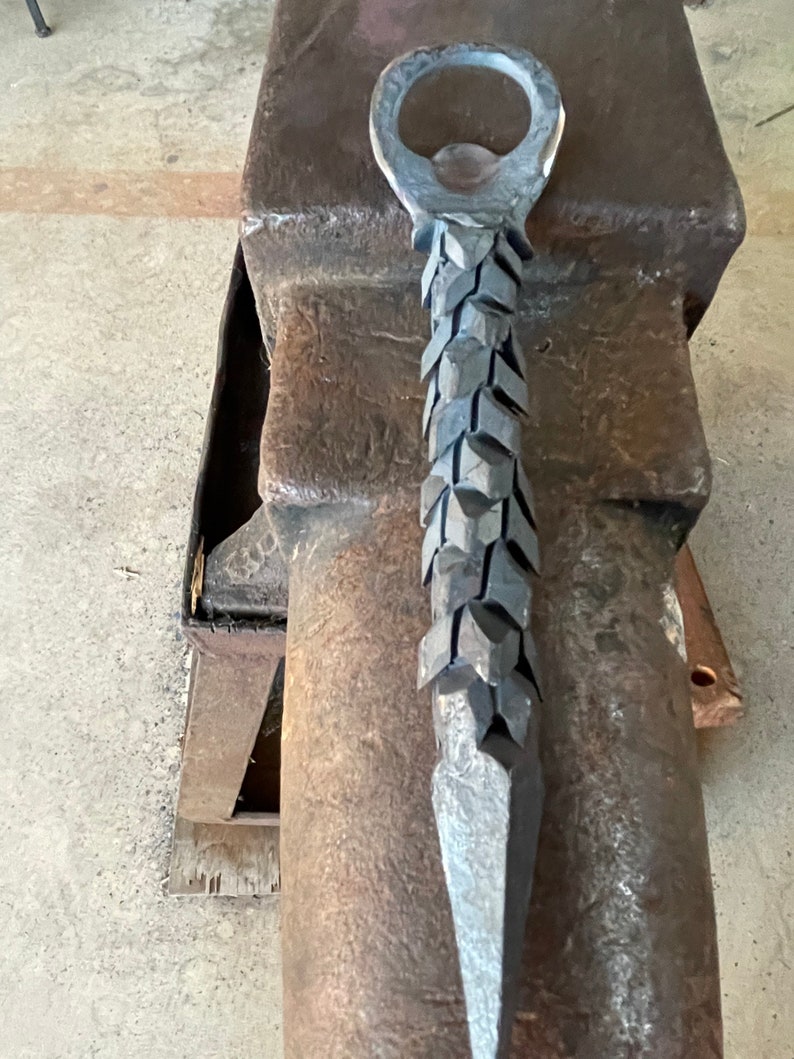 May include: A metal bottle opener with a unique, textured design. The opener is shaped like a spear with a ring at the top for opening bottles.