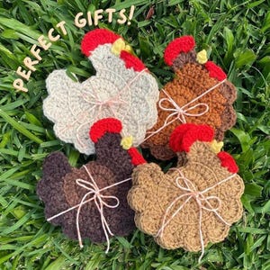 Op de afbeelding: Vier gehaakte onderzetters in de vorm van een kip in wit, donkerbruin, lichtbruin en beige, elk met een rode kam, gele snavel en een dun wit touwtje gestrikt tot een strik. De tekst PERFECT GIFTS! is zichtbaar in de linkerbovenhoek.