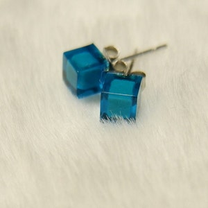以下が含まれることがあります： 銀のポストが付いた青い立方体のイヤリングのペア。イヤリングはガラス製で、クリアでファセットカットのデザインです。