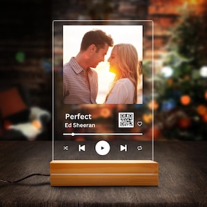 Pode incluir: Um leitor de música de acrílico transparente com uma base de madeira. O leitor apresenta uma foto de um casal, o título da música "Perfect" de Ed Sheeran, um código QR e controles de reprodução.