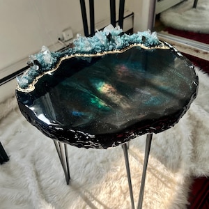 May include: A round side table with a dark, glossy resin top featuring swirling black, green, and iridescent hues. The edge is accented with textured, crystalline mineral formations in shades of teal and clear quartz, supported by three thin black metal legs.