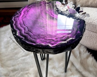 May include: A round side table with a purple and black agate design. The table has a glossy finish and is supported by three black metal legs. The top is adorned with crystals. The table is sitting on a white fluffy rug.
