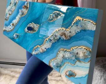 May include: A rectangular, abstract resin art piece with a vibrant ocean theme. The artwork features layers of turquoise and blue resin, accented with gold leaf and embedded crystals. The piece has a glossy finish and a unique, textured appearance.