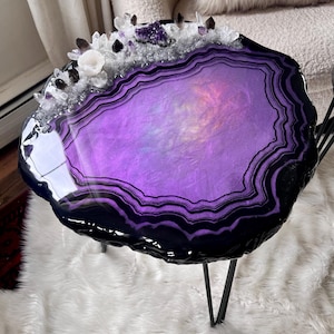 May include: A round side table with a glossy, irregular edge featuring a purple and black agate-inspired resin surface. The top is decorated with a cluster of clear, white, and dark purple crystals along one side, supported by thin black metal legs.