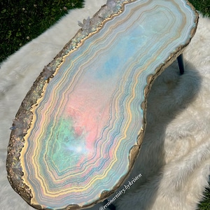 May include: A large, oval-shaped resin table with a light blue, pink, and green agate design. The table has a gold-colored edge and is decorated with clear quartz crystals. The table is sitting on a white fluffy rug. The text "@resinartistry_bykristen" is visible in the lower right corner of the table.
