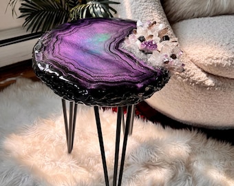 May include: A round side table featuring a purple and black agate-inspired resin top with a cluster of clear and purple quartz crystals embedded on one edge, supported by three thin black metal hairpin legs.