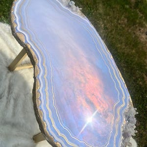 May include: An oval side table with a surface resembling a blue agate slice, featuring concentric rings of light blue, white, and gold. The edges are textured with gold paint and embedded clear crystals. The table stands on gold-colored legs.