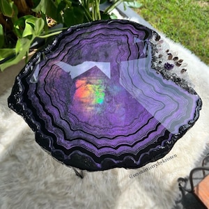 May include: A purple and black agate slice table top with a rainbow iridescent effect and clear resin. The table top is set on a black metal stand and has clear quartz crystals embedded in the resin around the edge. The table top is on a white fluffy rug. The text "@resinartistrybykristen" is in the lower right corner of the image.