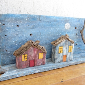 以下が含まれることがあります： 青い背景に白い星と月が描かれた、窓とドアが塗られた小さな木製の家が2つあります。家は流木で作られており、素朴で手作り感のある外観です。