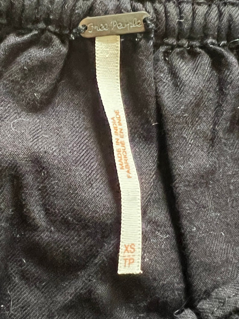 Może przedstawiać: Czarna metka odzieżowa Free People z białym tekstem, kt&oacute;ry brzmi "Made in India / Fabriqu&eacute; en Inde" i "XS TP".