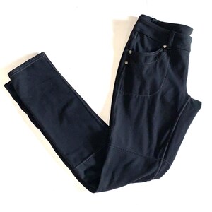 May include: A pair of black pants featuring a slim-fit design with a high waistband, belt loops, and two front pockets accented with small silver-toned metal rivets. The pants include visible stitching along the seams and a structured knee panel.