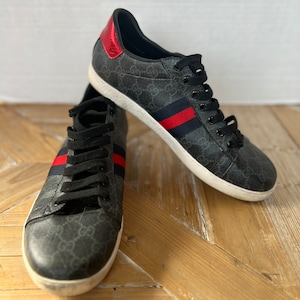 May include: A pair of black sneakers with a repeating pattern and red and blue stripes. The sneakers have black laces, a white sole, and a red accent on the heel. The shoes are displayed on a wooden surface.