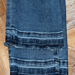 May include: Close-up of the bottom hem of a pair of dark blue denim jeans with a raw edge and a double stitched seam.