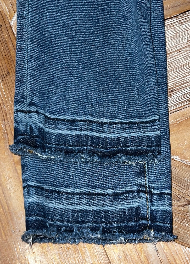 May include: Close-up of the bottom hem of a pair of dark blue denim jeans with a raw edge and a double stitched seam.