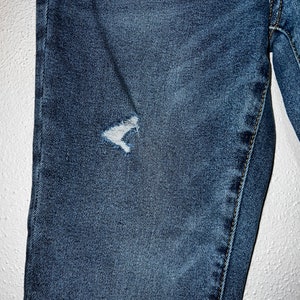 May include: Close-up of a pair of blue denim jeans with a small distressed area on the left thigh.