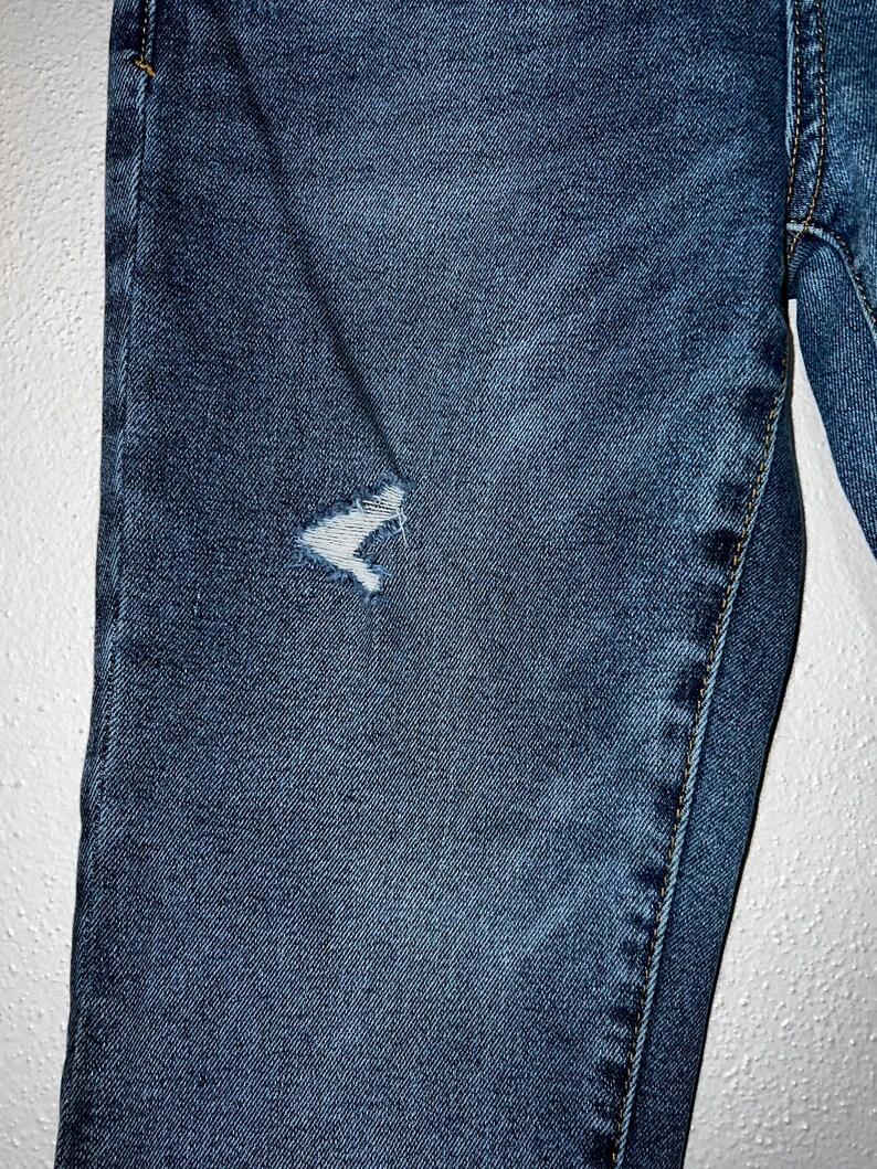 May include: Close-up of a pair of blue denim jeans with a small distressed area on the left thigh.
