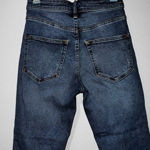 May include: A pair of dark blue denim jeans hanging on a clothes hanger. The jeans have a high waist and a slim fit.