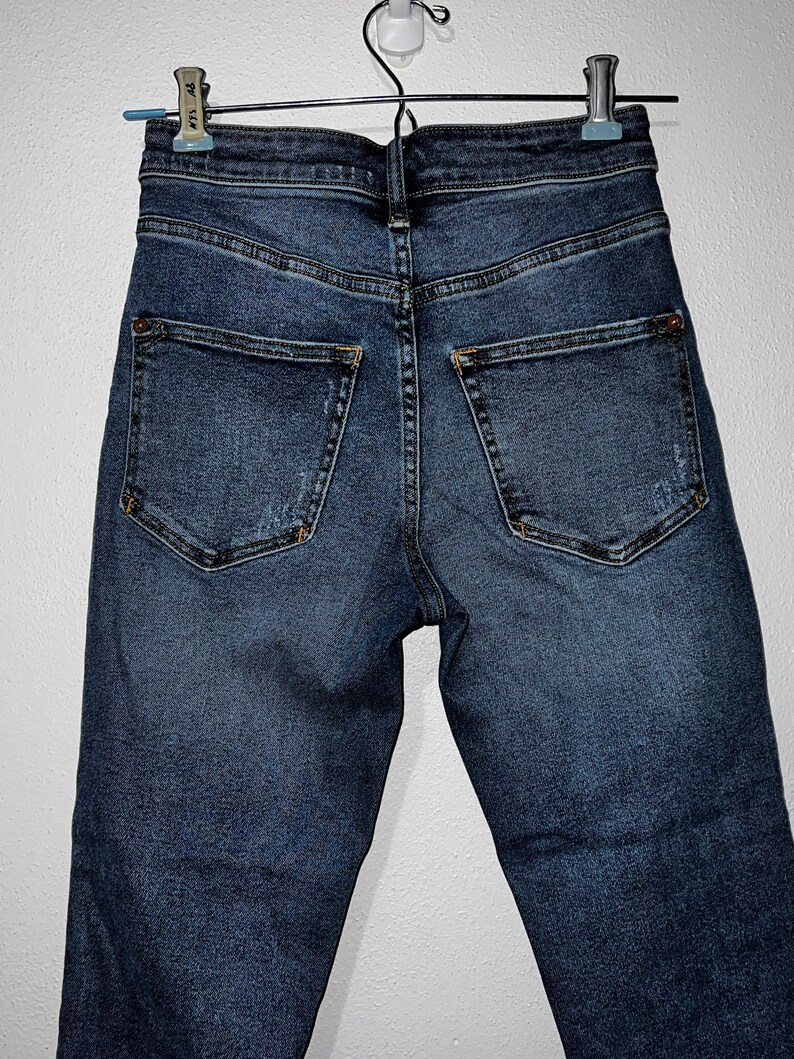 May include: A pair of dark blue denim jeans hanging on a clothes hanger. The jeans have a high waist and a slim fit.
