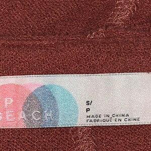 Op de afbeelding: Wit etiket met roze en blauwe cirkels en de tekst "FP BEACH" in zwart. Het etiket heeft ook de tekst "S/P" en "MADE IN CHINA FABRIQUE EN CHINE" in zwart.