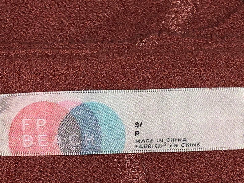 Op de afbeelding: Wit etiket met roze en blauwe cirkels en de tekst "FP BEACH" in zwart. Het etiket heeft ook de tekst "S/P" en "MADE IN CHINA FABRIQUE EN CHINE" in zwart.